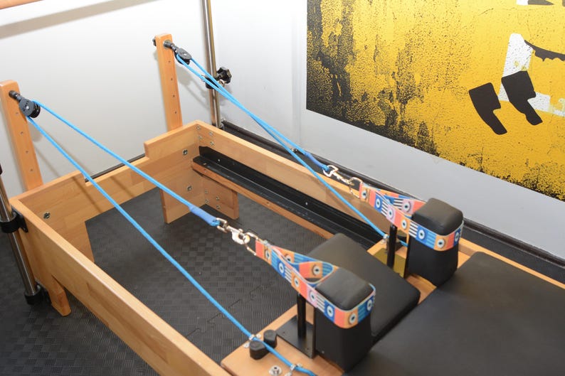 May include: A wooden Pilates reformer with blue straps and a black padded seat. The straps are attached to pulleys and have colorful patterned bands.