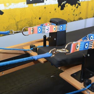 May include: Two blue ropes with black clips attached to a black and wooden pilates reformer. The ropes are connected to colorful straps with a pattern of eyes.