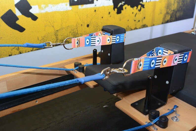 May include: Two blue ropes with black clips attached to a black and wooden pilates reformer. The ropes are connected to colorful straps with a pattern of eyes.