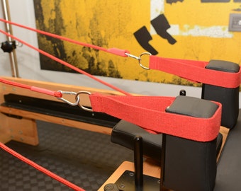 May include: Two red straps with metal clips attached to black padded blocks on a wooden pilates reformer.