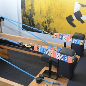 May include: A wooden Pilates reformer with two blue straps attached to black padded headrests. The straps have a colorful eye pattern.