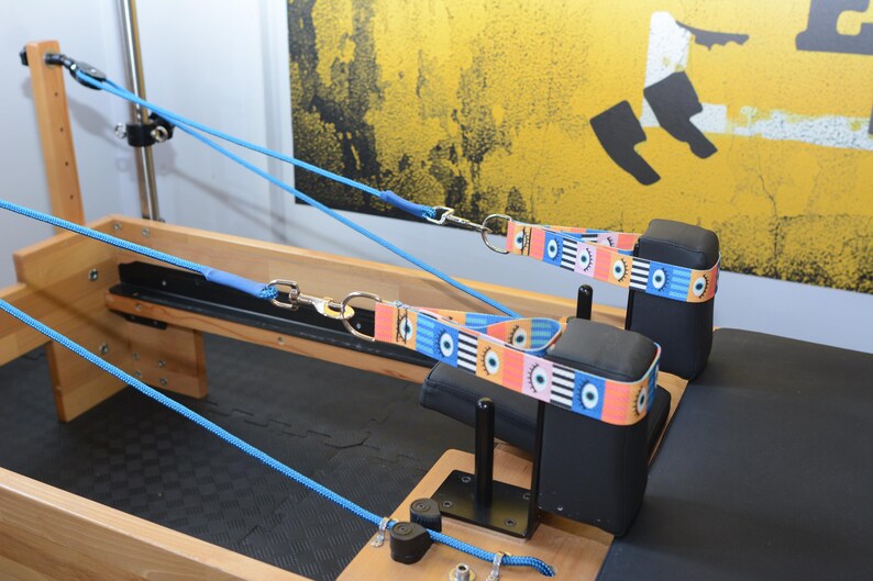 May include: A wooden Pilates reformer with two blue straps attached to black padded headrests. The straps have a colorful eye pattern.