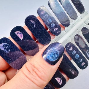 May include: Nail art stickers featuring a celestial design. The stickers display phases of the moon and planets against a dark blue and purple gradient background. The nail art stickers are on a clear plastic sheet.