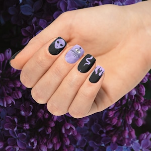 May include: Close-up of a hand with black nail polish and various designs. The nails feature an alien head, an eye with a star, a snake, and a wavy design.