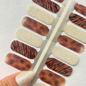 May include: Nail art strips featuring a mix of designs. Some strips have a tiger stripe pattern in brown and black, others are a solid gold glitter, and some have a gradient of brown and tan. The nail strips are on a white background.