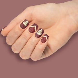 May include: A hand with burgundy nail polish with gold glitter and a gold Christmas tree design on each nail.