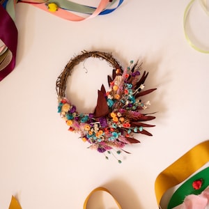 May include: A dried flower wreath with a variety of colors, including purple, pink, orange, and blue. The wreath is made of twigs and is decorated with dried flowers and leaves. The wreath is surrounded by colorful ribbons.