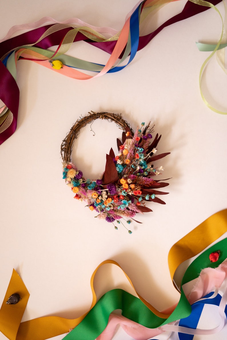 May include: A dried flower wreath with a variety of colors, including purple, pink, orange, and blue. The wreath is made of twigs and is decorated with dried flowers and leaves. The wreath is surrounded by colorful ribbons.