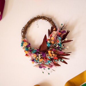 May include: A dried flower wreath with a variety of colors, including purple, pink, orange, and blue. The wreath is made of twigs and is decorated with dried flowers and leaves.
