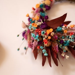 May include: A dried flower wreath with a variety of colors, including orange, pink, purple, blue, and green. The wreath is made with dried flowers and leaves, and it is perfect for adding a touch of natural beauty to any home.
