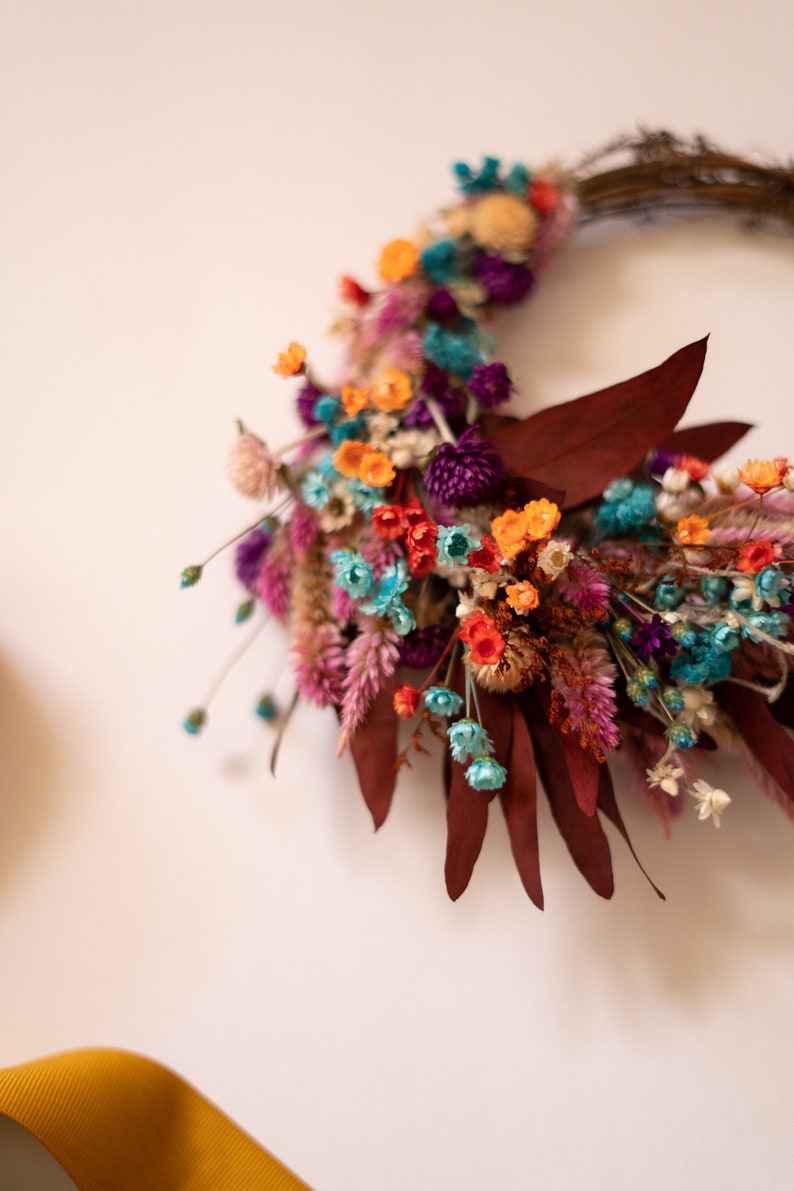 May include: A dried flower wreath with a variety of colors, including orange, pink, purple, blue, and green. The wreath is made with dried flowers and leaves, and it is perfect for adding a touch of natural beauty to any home.