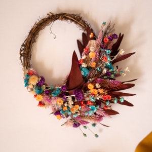 May include: A dried flower wreath with a variety of colors, including purple, orange, pink, and blue. The wreath is made of twigs and has a rustic look.