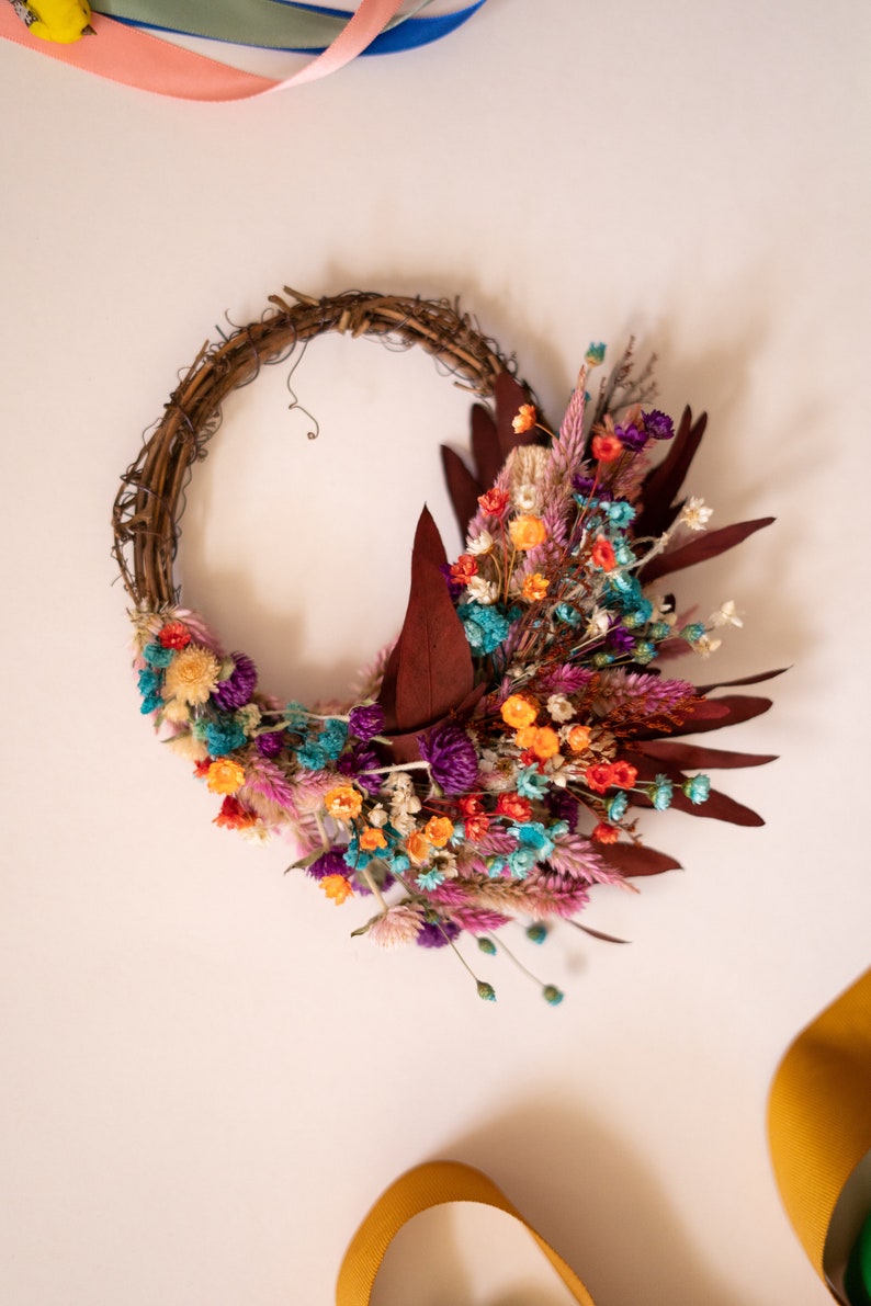 May include: A dried flower wreath with a variety of colors, including purple, orange, pink, and blue. The wreath is made of twigs and has a rustic look.