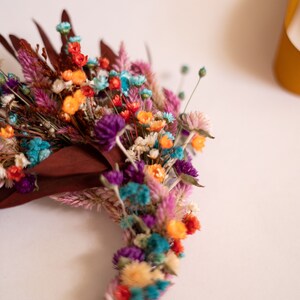 May include: A colorful dried flower wreath with a variety of colors including pink, purple, blue, orange, and yellow. The wreath is made with a natural twig base.