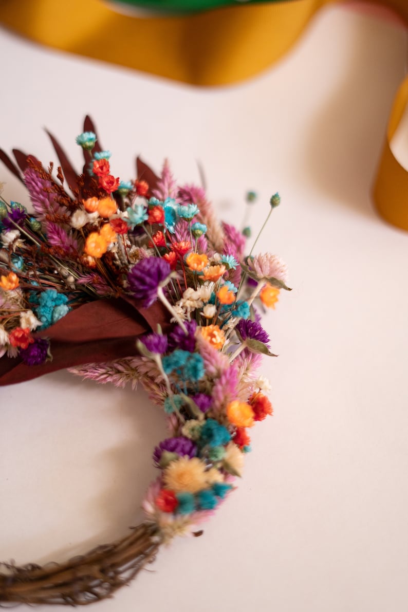 May include: A colorful dried flower wreath with a variety of colors including pink, purple, blue, orange, and yellow. The wreath is made with a natural twig base.