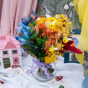 May include: A vibrant bouquet of dried flowers in a clear glass vase, featuring a rainbow of colors including blue, green, orange, and yellow. The arrangement is adorned with small bee decorations and tied with a purple ribbon. A whimsical floral display.