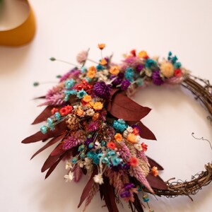 May include: A dried flower wreath with a variety of colors, including pink, purple, orange, blue, and white. The wreath is made with natural materials and has a rustic look.