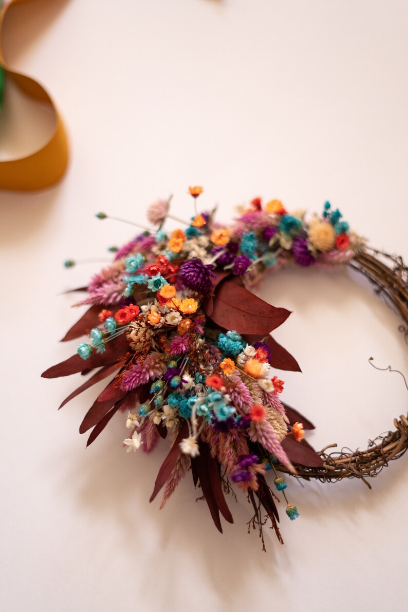 May include: A dried flower wreath with a variety of colors, including pink, purple, orange, blue, and white. The wreath is made with natural materials and has a rustic look.