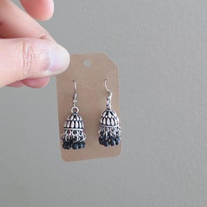 May include: A pair of silver-toned jhumka earrings featuring a bell-shaped design with intricate embossed patterns and small, dangling black beads at the base. The earrings are attached to a rectangular brown cardboard display tag with a hole at the top.