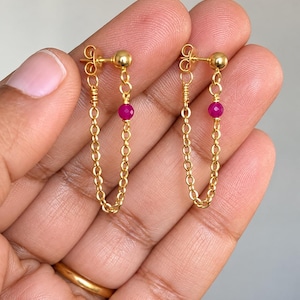 May include: A pair of gold chain earrings featuring a small, round, faceted fuchsia-colored bead on each chain. The earrings are designed as a loop, connecting a gold stud post to a gold link chain, creating a delicate and dangling drop effect.