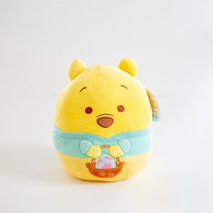 May include: Yellow plush Winnie the Pooh holding a basket with an Easter egg. He is wearing a blue and white shirt with a heart on the front.