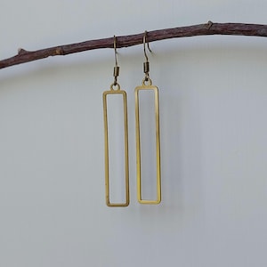 May include: A pair of gold-coloured rectangular earrings suspended from a brown branch. The earrings feature a simple, open frame design with a hook closure. The background is a plain, off-white colour.