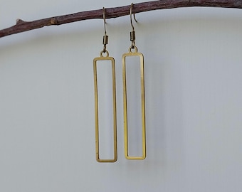 Puede incluir: Un par de pendientes rectangulares dorados colgando de una rama marrón. Los pendientes tienen un diseño de marco abierto simple con cierre de gancho. El fondo es de color blanquecino.