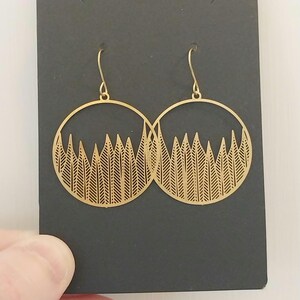Puede incluir: Pendientes de aro dorados con diseño de bosque. Cada pendiente presenta un marco circular con un patrón recortado de árboles o follaje estilizados. Los pendientes se muestran en una tarjeta negra.