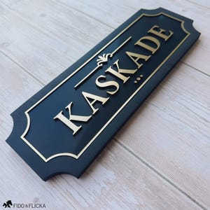 May include: A rectangular dark blue sign with rounded corners and a gold-coloured border. The sign features the word KASKADE in raised gold-coloured capital letters, with a decorative gold-coloured flourish above the text and three small dots below.