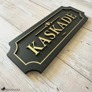 Puede incluir: Letrero rectangular negro con la palabra "KASKADE" en letras doradas. El letrero tiene un borde dorado y elementos decorativos. Probablemente para una casa o negocio.
