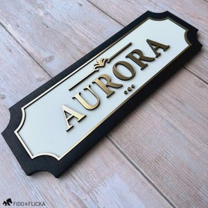 May include: A rectangular sign with the name "AURORA" in gold lettering. The sign has a white centre, gold trim, and a black border. Decorative elements include a flourish above the name and three dots below.
