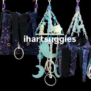 May include: Two dark purple galaxy-print fabric pouches with black interior lining and metal grommets, hanging alongside a central mobile featuring light blue plastic crescent moons, stars, and white plastic chains. The text ihartsuggies is visible.