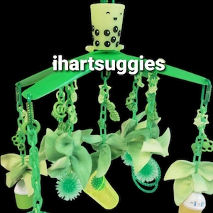 May include: A green and yellow baby mobile with a boba tea cup at the top. The mobile has a variety of green and yellow toys hanging from it, including a bow, a bottle, and a star.