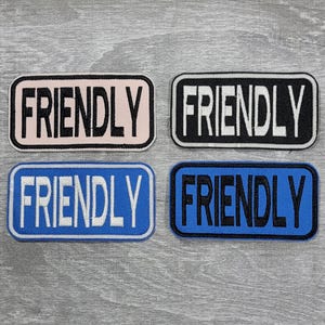 May include: Four embroidered patches with the word "FRIENDLY" in different colors and fonts. The patches are on a gray wooden background.