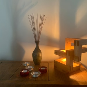 Puede incluir: Una lámpara de madera con un diseño geométrico, iluminada por una vela en su interior. La lámpara está sobre una mesa de madera con un jarrón de ramas secas y varias velas de té.