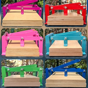 May include: A selection of brightly coloured tortilla presses. Each press has a wooden base and a metal frame in various colours, including green, yellow, orange, pink, red, blue, and teal. The presses are for making tortillas.