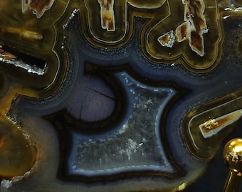 Pode incluir: Close-up de uma fatia de geodo de ágata polida com um centro azul escuro e roxo rodeado por bandas concêntricas brancas, marrons e amarelas. A geoda está montada em um suporte de metal dourado.