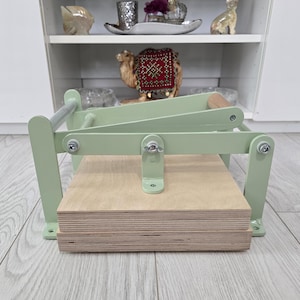 May include: A light green book press with a natural wood base. The press has a metal frame and a wooden handle. The press is designed for pressing books or art. The press is on a light wood floor. The press measures approximately 30 cm in length.