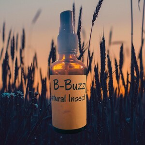 May include: Amber glass spray bottle with a black cap and a yellow label. The label reads "B-Buzz Natural Insect". The bottle is set against a backdrop of tall grass and a sunset.