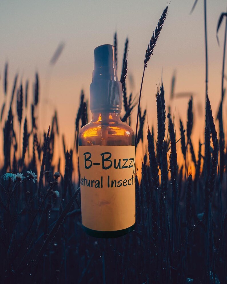 May include: Amber glass spray bottle with a black cap and a yellow label. The label reads "B-Buzz Natural Insect". The bottle is set against a backdrop of tall grass and a sunset.