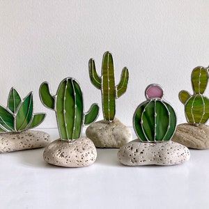 May include: Five stained glass cactus figurines on small grey rocks. The cacti are green and have different shapes and sizes. One cactus has a pink flower.