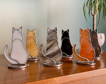 May include: Six stained glass cat figurines in various colors, including white, orange, brown, and black. The cats are all facing the same direction and are sitting on a wooden surface.