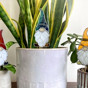 May include: Three stained glass gnomes with white beards and different colored hats. The gnomes are placed in potted plants. The gnome on the left has a red hat, the gnome in the middle has a blue hat, and the gnome on the right has a yellow hat.