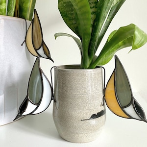 May include: Three stained glass birds, one white and gray, one yellow and gray, and one brown and gray, are hanging from the rim of a gray ceramic pot. A green plant is growing in the pot.
