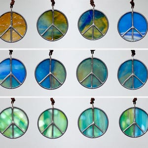 May include: A collection of round, stained glass peace sign pendants in various colors. Each pendant features a silver-colored metal frame and a brown leather cord for hanging. The glass colors include shades of blue, green, and yellow.