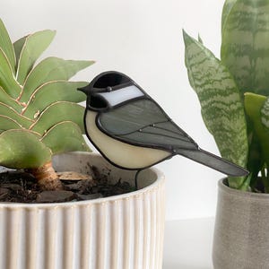 May include: A stained glass bird decoration in a garden setting. The bird has a black head, white accents, and gray wings and body. It is perched on a wire stake, placed in a white ceramic pot with a plant. Other plants are in the background.