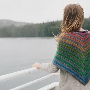May include: A person wearing a gray sweater and a colorful, striped shawl with a rainbow of colors. The shawl is draped over the shoulders, with the person looking out at a body of water and a forested shoreline.
