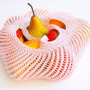 Puede incluir: Una cesta de frutas de punto rosa claro llena de una pera amarilla, fresas rojas y rodajas de naranja. La cesta tiene un diseño de tejido abierto único, que permite ver las frutas coloridas.