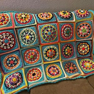 May include: A colorful, handmade crochet blanket with a patchwork design. The blanket features square motifs in various colors, including turquoise, orange, yellow, and pink. Each square has a central floral design. The blanket is draped over a brown sofa.