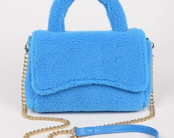 Könnte beinhalten: Eine kleine blaue Handtasche aus Kunstfell mit einer goldenen Kettenriemenschlaufe und einem oberen Henkel. Die Tasche hat einen Klappenverschluss und eine weiche, flauschige Textur.